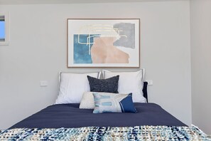 A bedroom with a bed, pillows, a blanket, and a framed abstract painting on the wall.