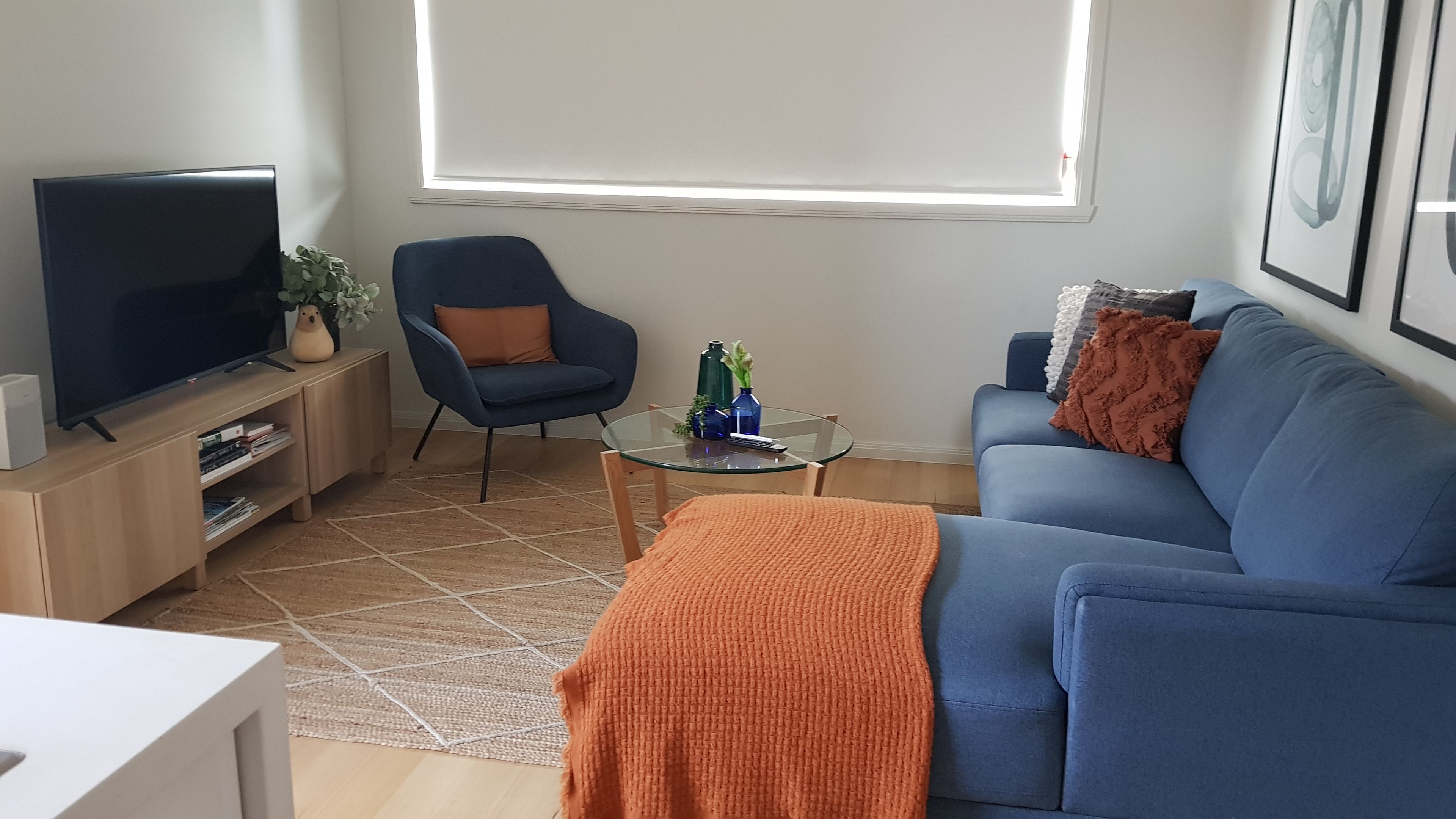 A living room with a television, a blue sofa, a blue armchair, a glass coffee table with decorative items, and a large window.