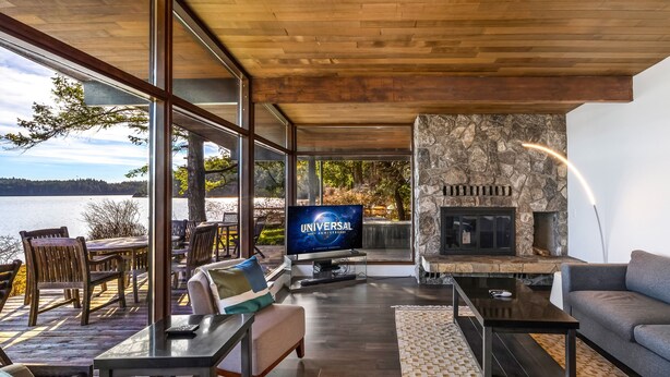 A modern living room with a stone fireplace, a television displaying 'Universal 100th Anniversary', and a view of a lake through large windows. - Waterfront Home! Newly Remodeled! (weow) (Friday harbor)