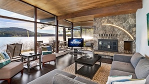 A modern living room with a stone fireplace, large windows overlooking a lake, and a television displaying 'Universal 50th Anniversary'. - Waterfront Home! Newly Remodeled! (weow) (Friday harbor)