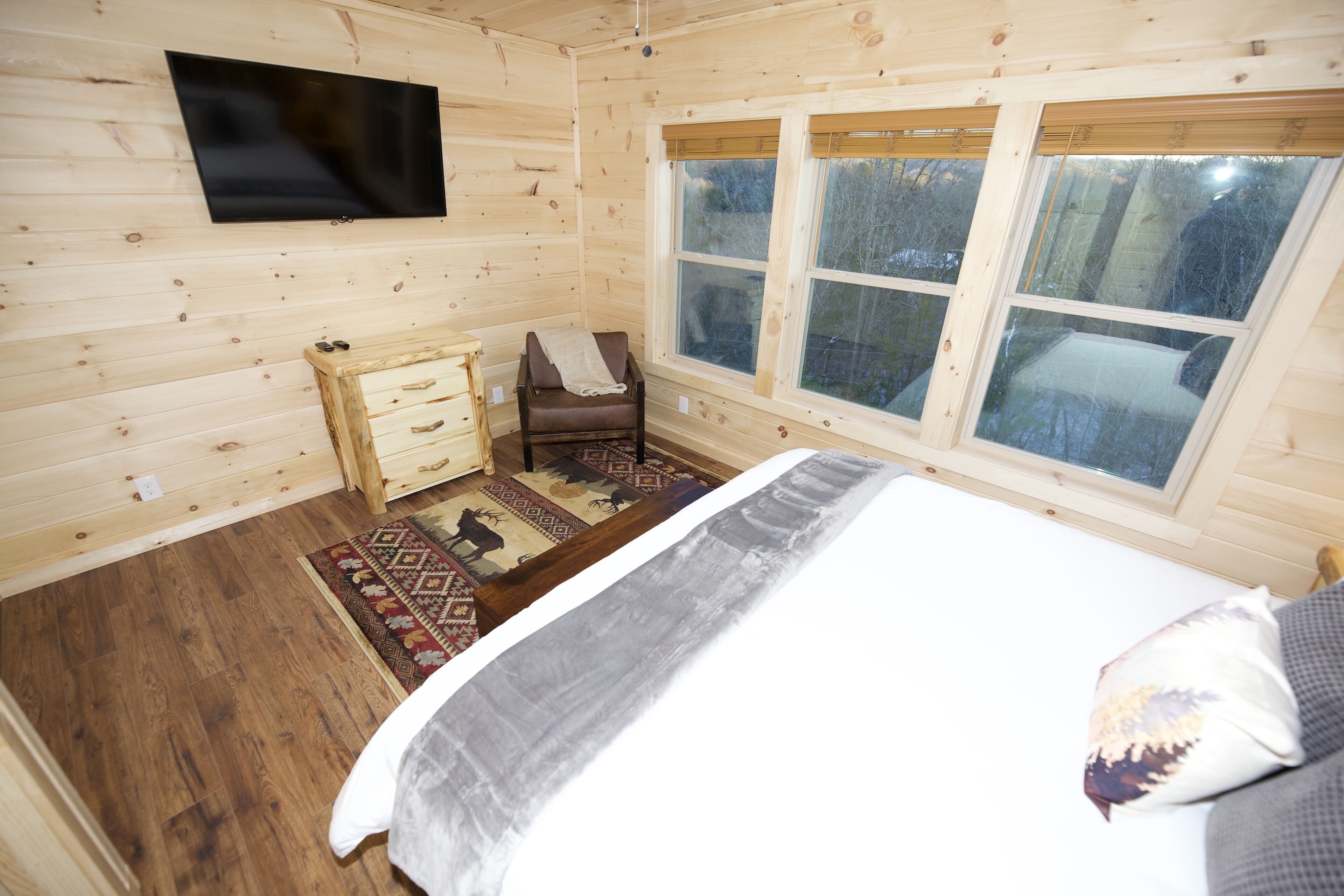 Een houten slaapkamer met een bed, een aan de muur bevestigde televisie en een raam met uitzicht op bomen.
