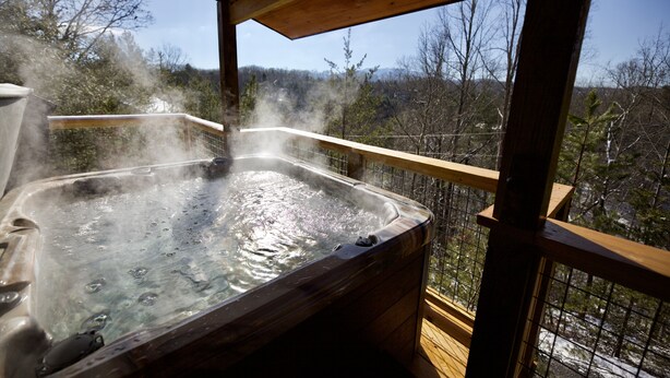 A hot tub with steam rising, surrounded by a wooden deck and a view of trees and hills. - Quiet Family Cabin on Private Mountain Top-Awesome Location! (Sevierville)