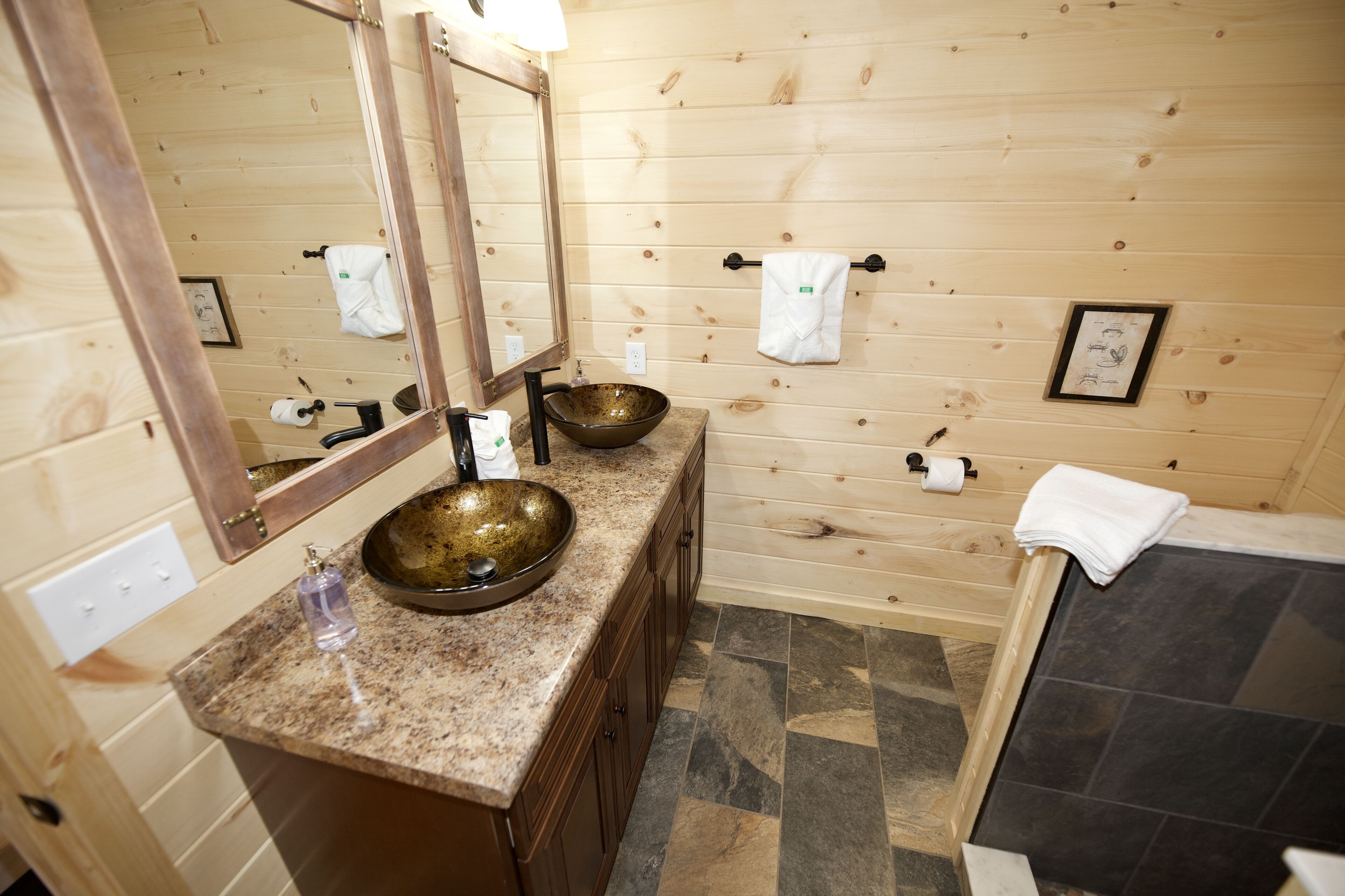 A wooden cabin-style bathroom with a large mirror, two sinks, a bathtub, and a tiled floor.