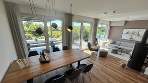 Een moderne woonkamer met een houten eettafel, zwarte stoelen, een grijze bank en een groot raam met uitzicht op groen en een zwembad.