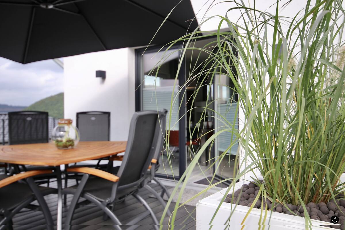 An outdoor dining area with a wooden table and chairs, a black umbrella, and a potted plant with tall green grass.
