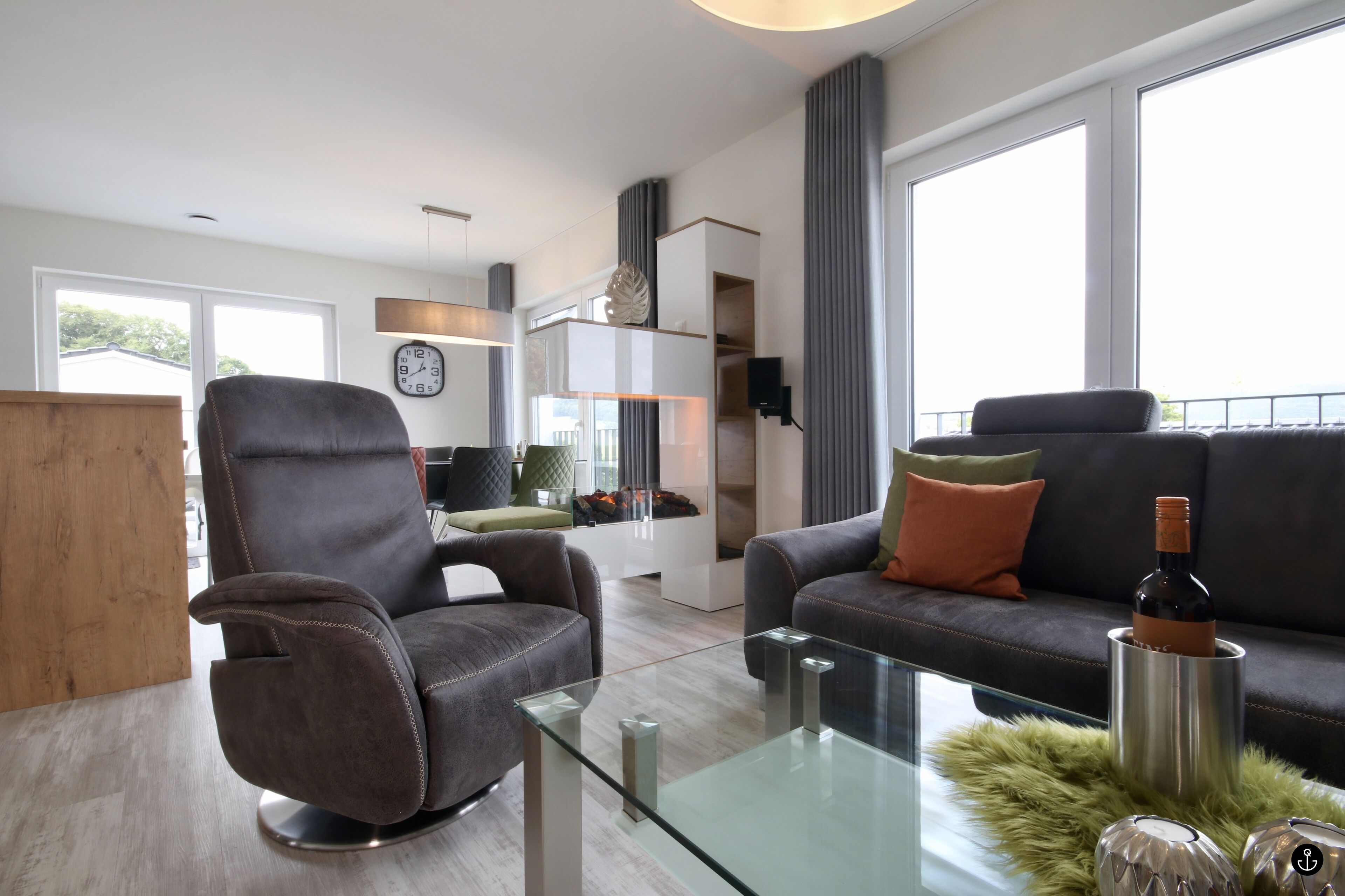Un salon moderne avec un fauteuil en cuir, une table basse en verre et un canapé agrémenté de coussins. On aperçoit une bouteille sur la table et une horloge au mur.