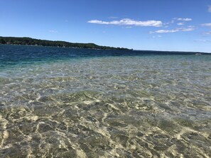 A clear view of a beach with shallow water, a distant shoreline with trees, and a boat in the distance.