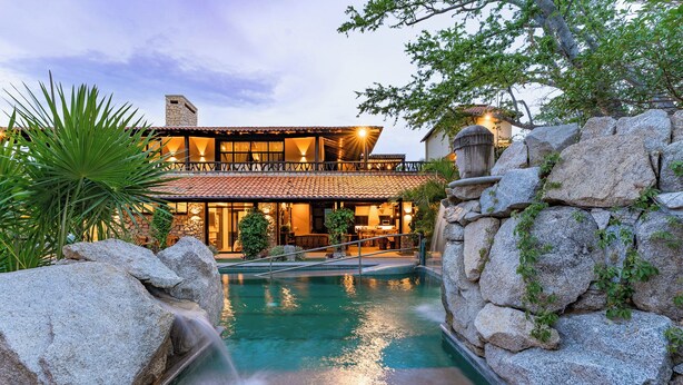 A pool with a stone waterfall, surrounded by large rocks and greenery, leading to a building with a tiled roof and large windows.