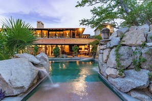 Une piscine avec une cascade en pierre, entourée de gros rochers et de verdure, qui mène à un bâtiment doté d’un toit en tuiles et de grandes fenêtres.