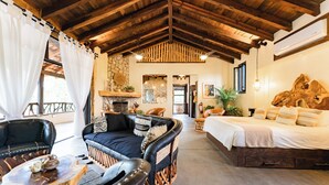 A spacious room with a high wooden ceiling, a stone fireplace, a bed with a large headboard, and a seating area with a coffee table and chairs.