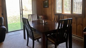 A wooden dining room with a large table and chairs, a hanging light fixture, and a view of the outdoors through a sliding door.
