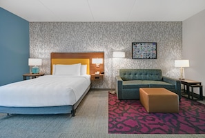 Una habitación de hotel con cama, sofá, sillón, una mesita pequeña y una obra de arte colgada en la pared.