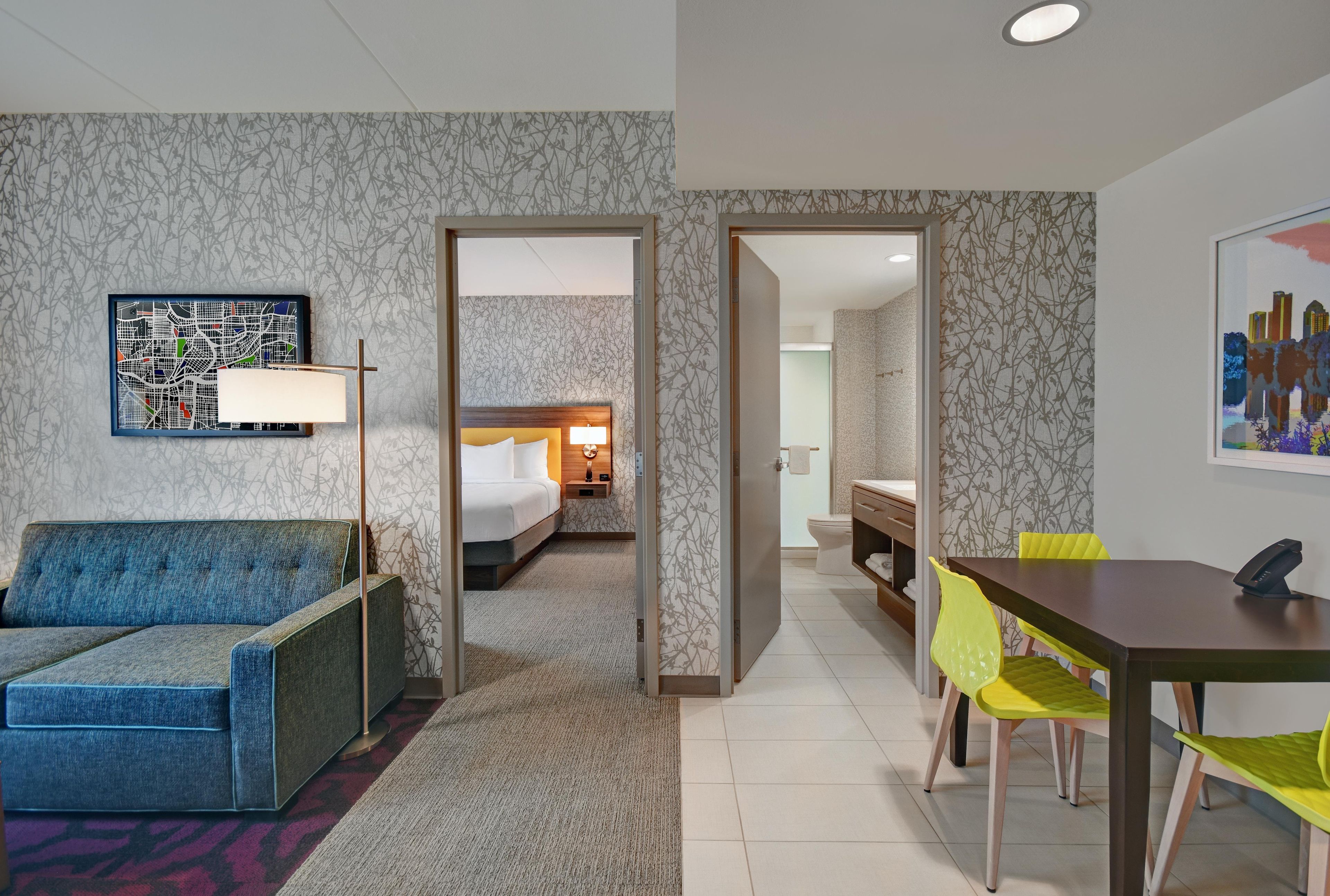 Habitación de hotel con cama, sofá, mesa de comedor con sillas y baño visible a través de una puerta abierta.