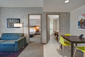 Habitación de hotel con cama, sofá, mesa de comedor con sillas y baño visible a través de una puerta abierta.
