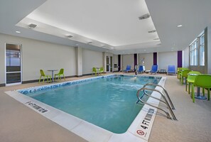 Una piscina interior con agua cristalina, rodeada de sillones de jardín verdes y azules, y un letrero que indica 'no saltos' en el borde de la piscina.