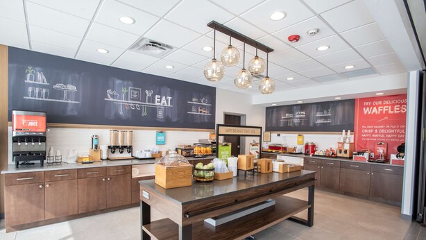 A modern cafe with a chalkboard menu, a juice station, and a waffle display. - Hampton Inn by Hilton Richwood Cincinnati South (Walton)