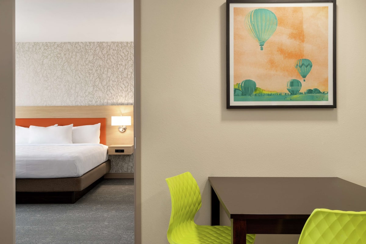 Une chambre d’hôtel avec un lit, une table de chevet, une lampe, un tableau encadré représentant des montgolfières et une table avec une chaise.