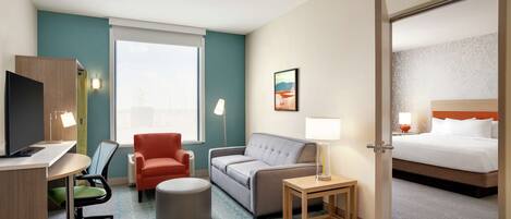 A hotel room with a desk, a grey sofa, a red armchair, a small ottoman, a bedside table with a lamp, and a bed with white bedding.