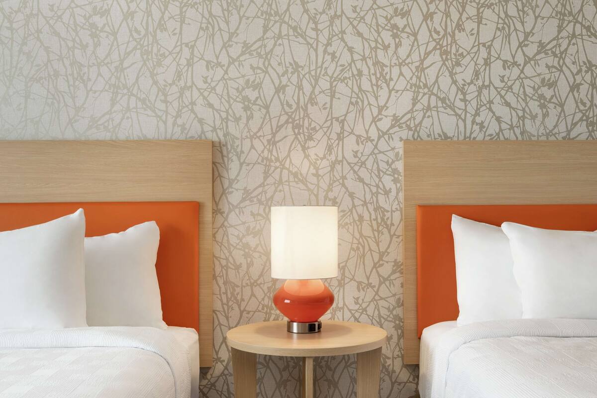 Une chambre d’hôtel avec deux lits, chacun doté d’une tête de lit orange, d’une table de chevet ronde en bois avec une lampe rouge, et d’un papier peint à motifs.