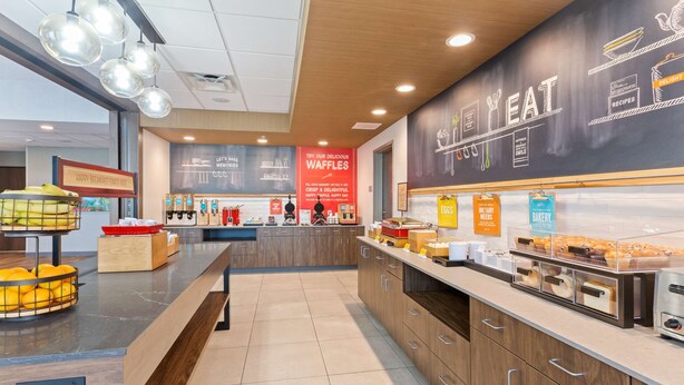 A modern kitchen area with a central counter, various food stations, and a chalkboard menu. - Hampton Inn & Suites Alachua I-75 (Alachua)