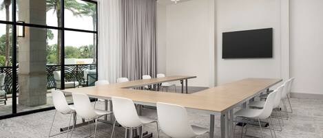A conference room with a long table, white chairs, a large window with a view of palm trees, and a flat-screen TV mounted on the wall.