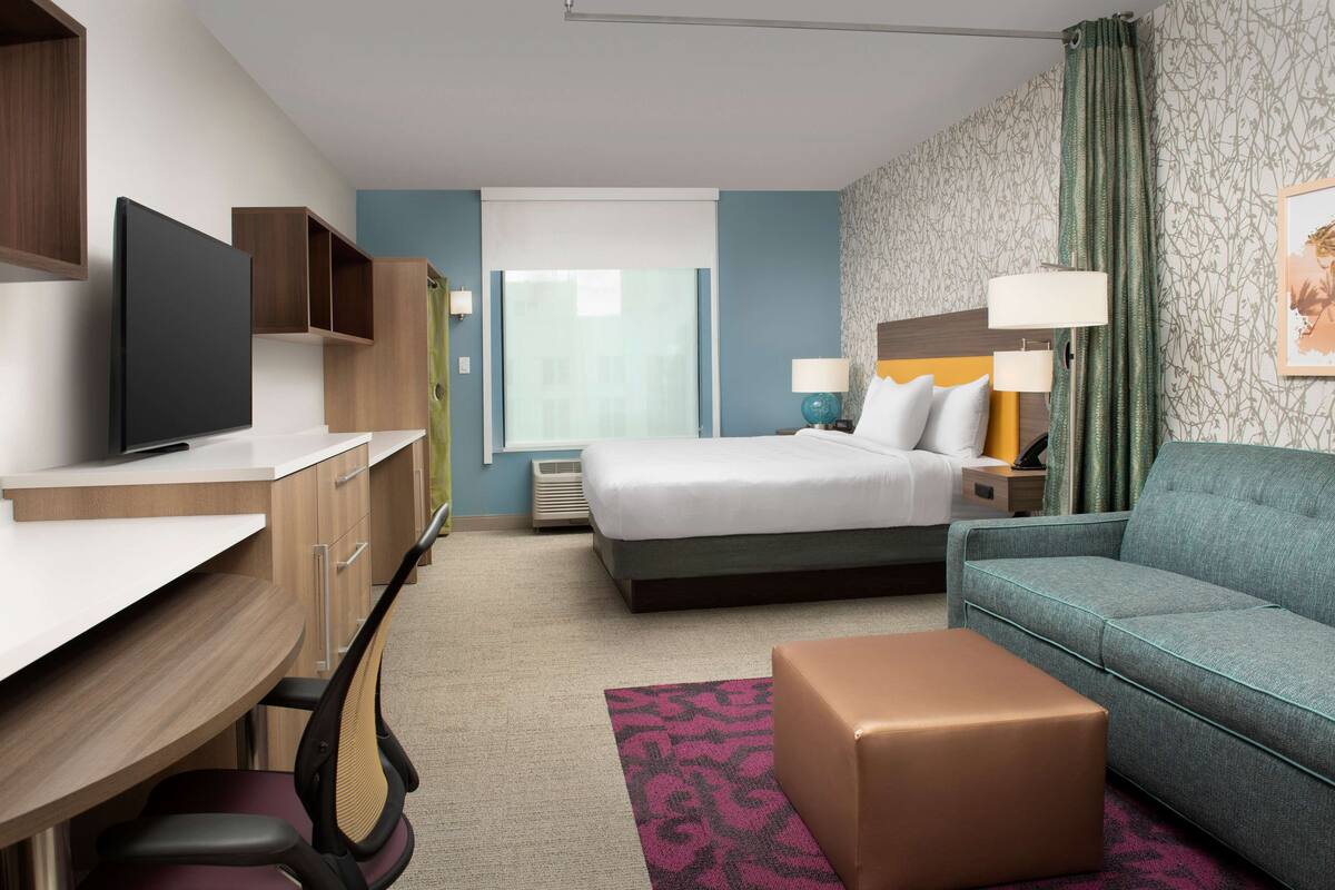 Une chambre d’hôtel comprenant un lit, un canapé, un bureau avec une chaise, une télévision et une fenêtre avec des rideaux.