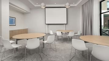 Une salle de conférence équipée de tables rondes, de chaises blanches, d’un écran de projection et d’une grande fenêtre avec des rideaux.