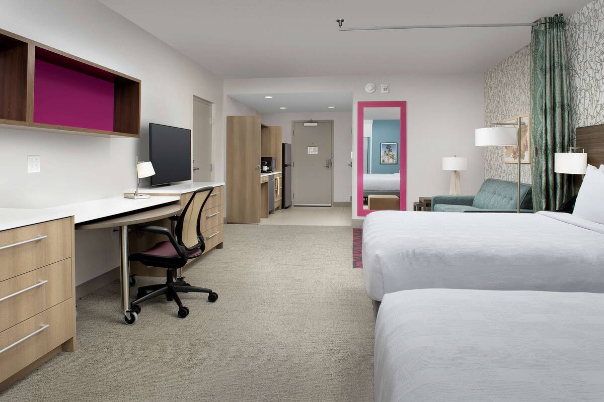 Une chambre d’hôtel équipée d’un bureau, de deux lits, d’une télévision et d’une salle de bain visible à travers une porte ouverte.