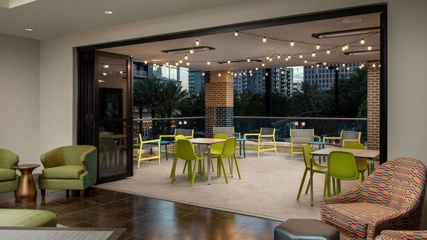 Un espace de restauration intérieur moderne, avec des chaises et des tables vertes, de grandes fenêtres offrant une vue sur la ville et des guirlandes lumineuses au plafond.