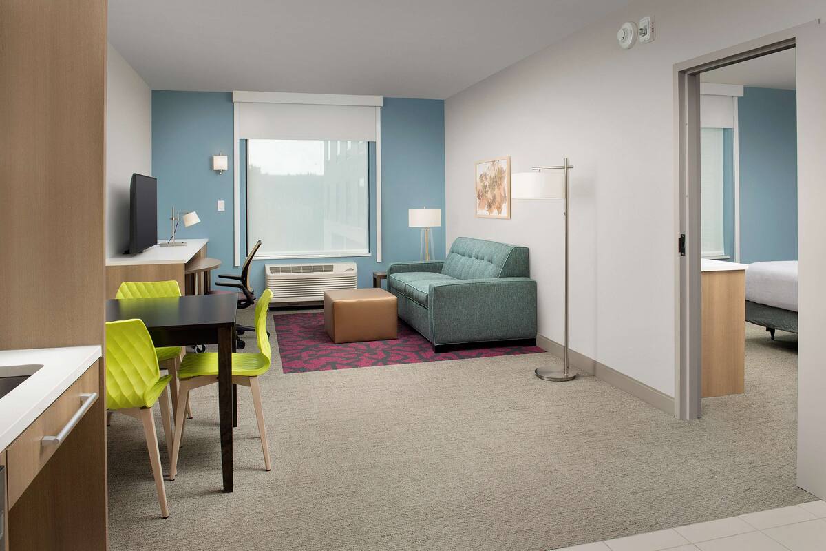 Une chambre d’hôtel avec un bureau, un canapé vert, un pouf marron et un lit.