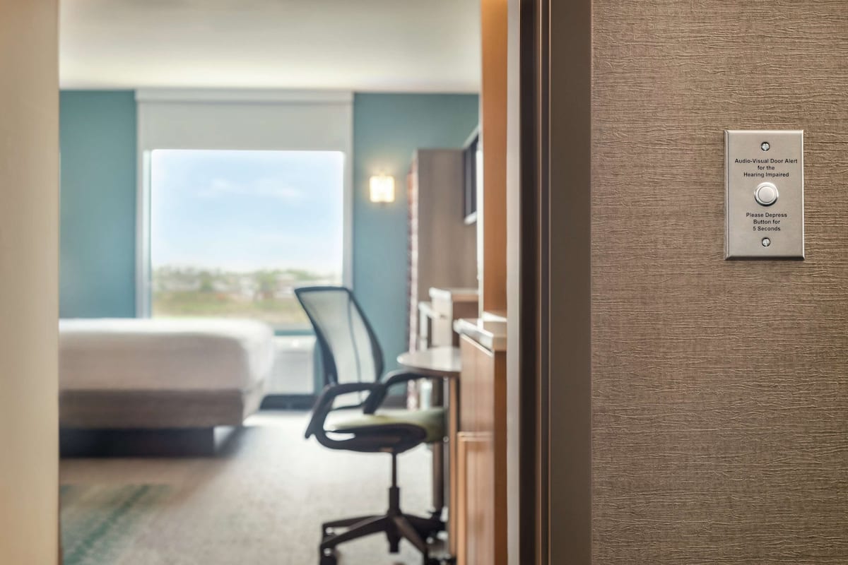 A hotel room with a bed, a desk, and a chair. There is a sign on the wall indicating an audio-visual door alert system.