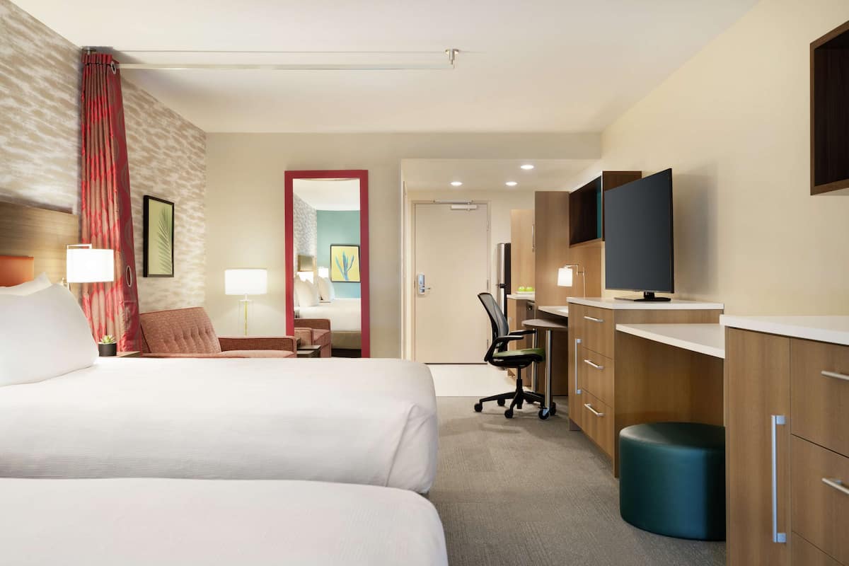A hotel room with two beds, a desk, a chair, a TV, and a bathroom visible through an open door.