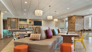 A modern living room with a sectional sofa, orange ottomans, a dining area, and a kitchen with a stone accent wall. - Home2 Suites by Hilton Mesa Longbow, AZ (Mesa)