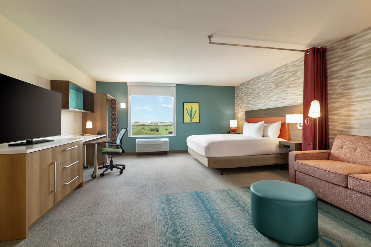 A hotel room with a bed, a desk with a TV, a chair, a sofa, a window with a view, and a lamp.