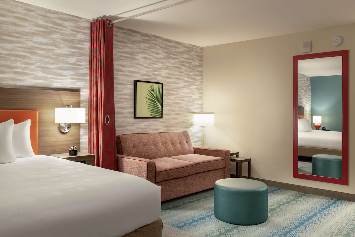 A hotel room with a large bed, a sofa, a small ottoman, a mirror, and a wall-mounted lamp.