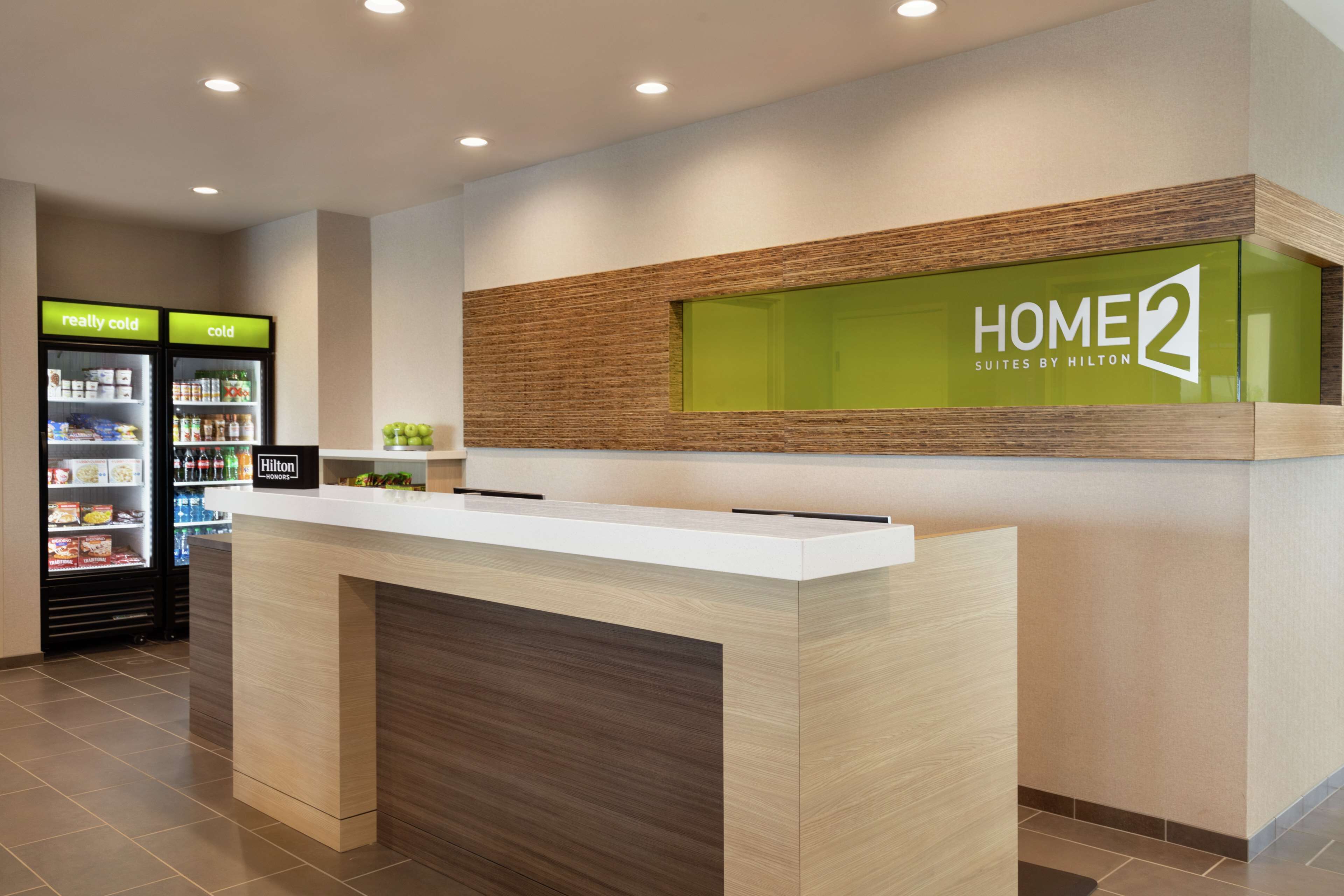Un comptoir d’accueil avec une enseigne indiquant « HOME2 SUITES BY HILTON » et un coin bar avec des étiquettes indiquant « très froid » et « froid ».