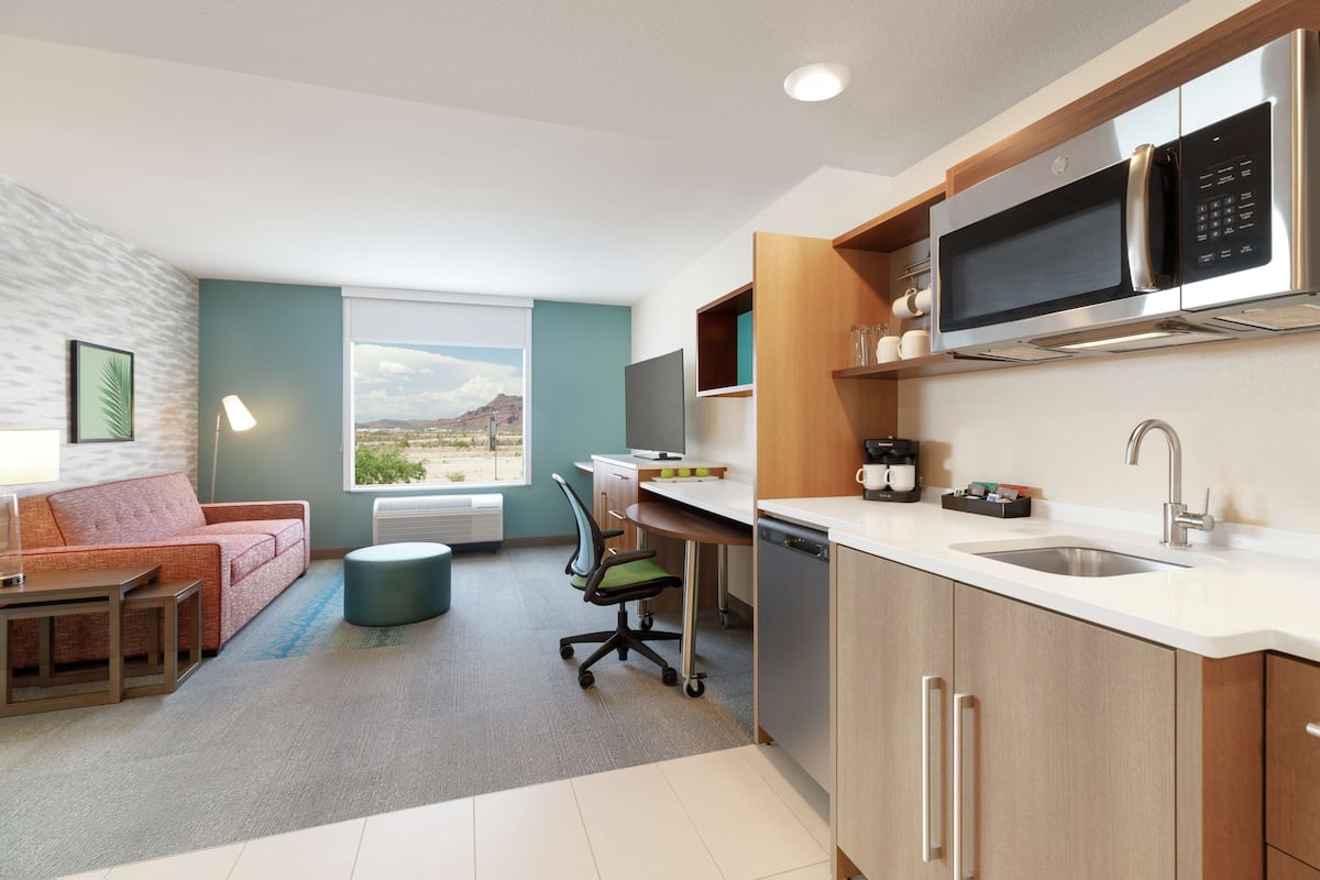 A modern kitchen with a microwave, sink, and dishwasher. There is a dining area with a table and chairs, and a sofa. The room has a large window with a view of the mountains.