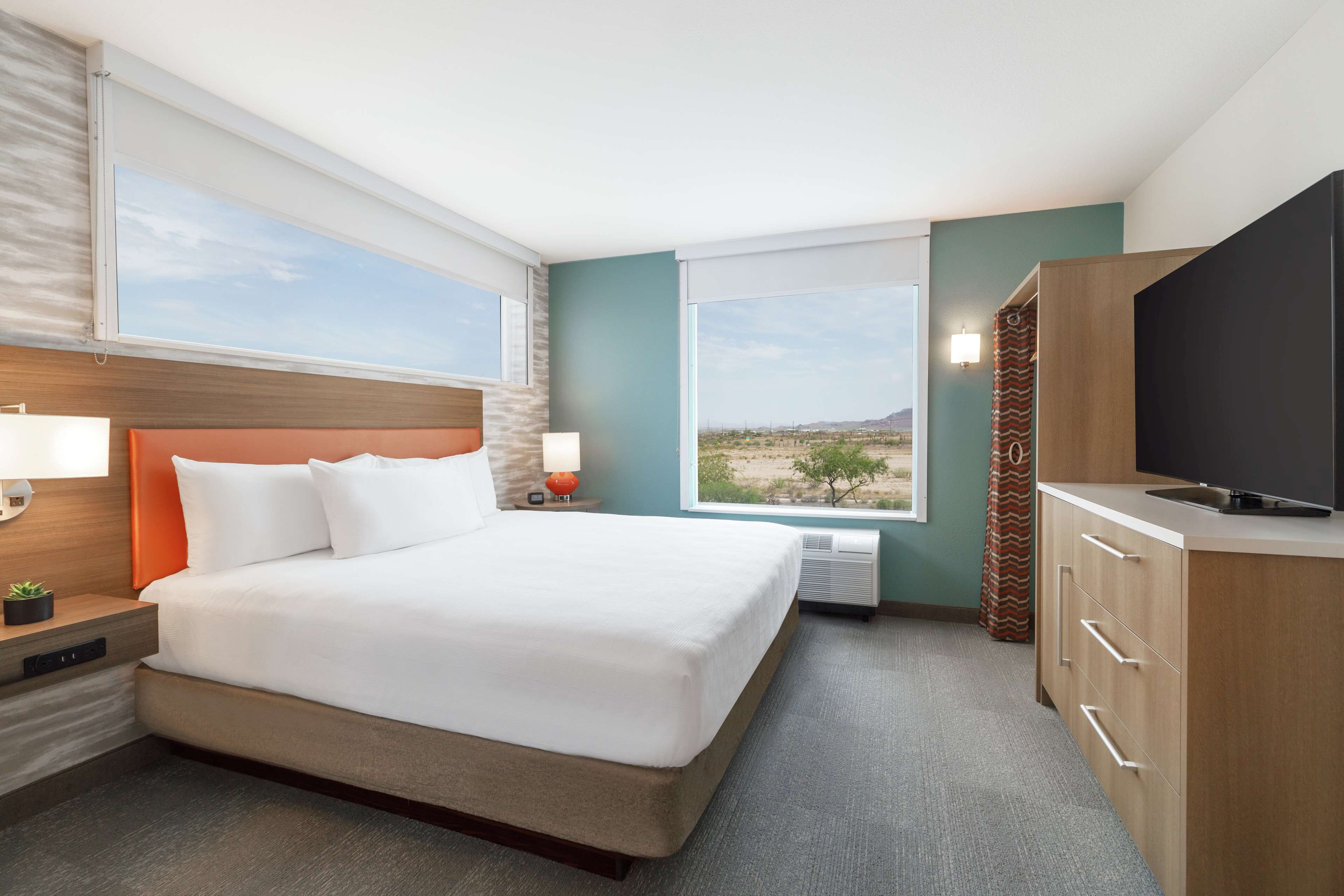 A hotel room with a large bed, a TV, a desk with a lamp, and a view of the desert.
