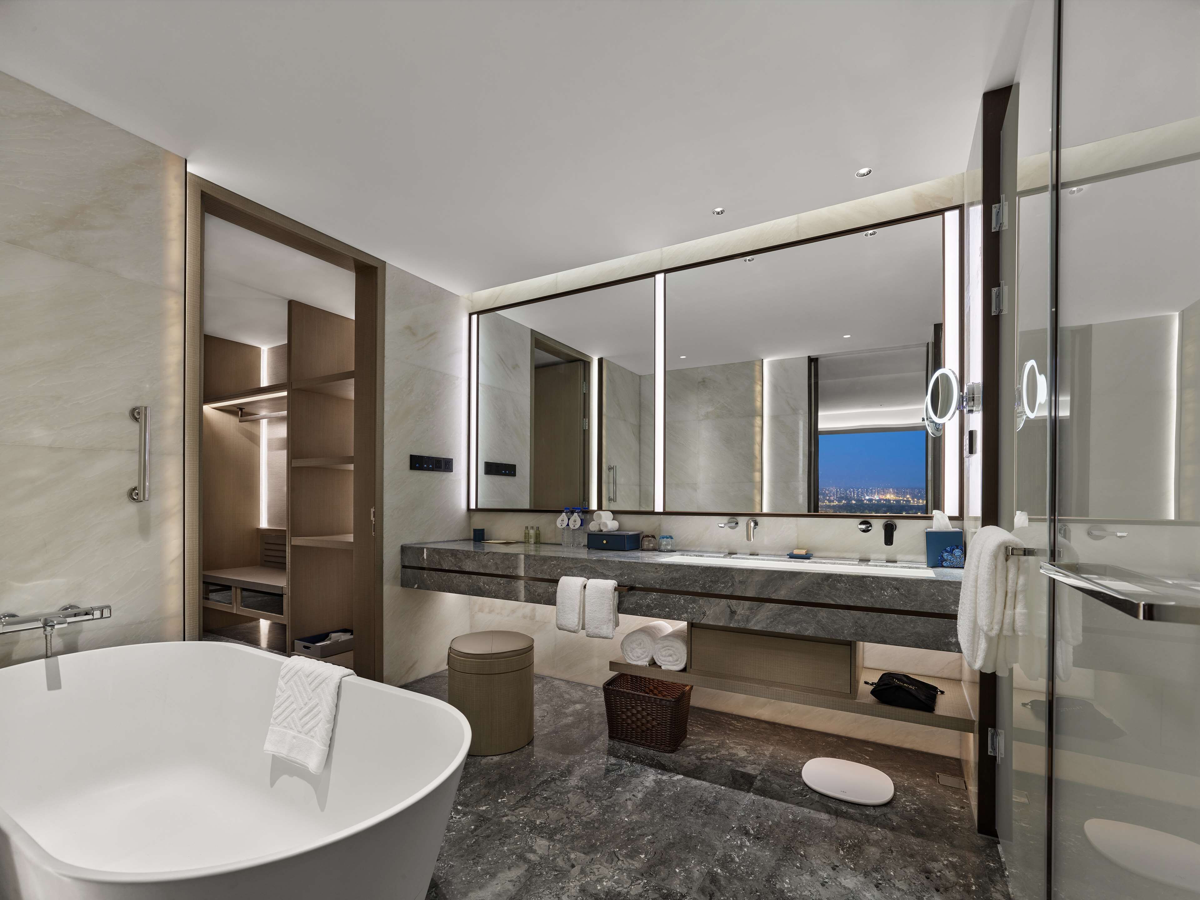 Un baño moderno con una bañera independiente, un espejo grande y vista a la ciudad de noche.