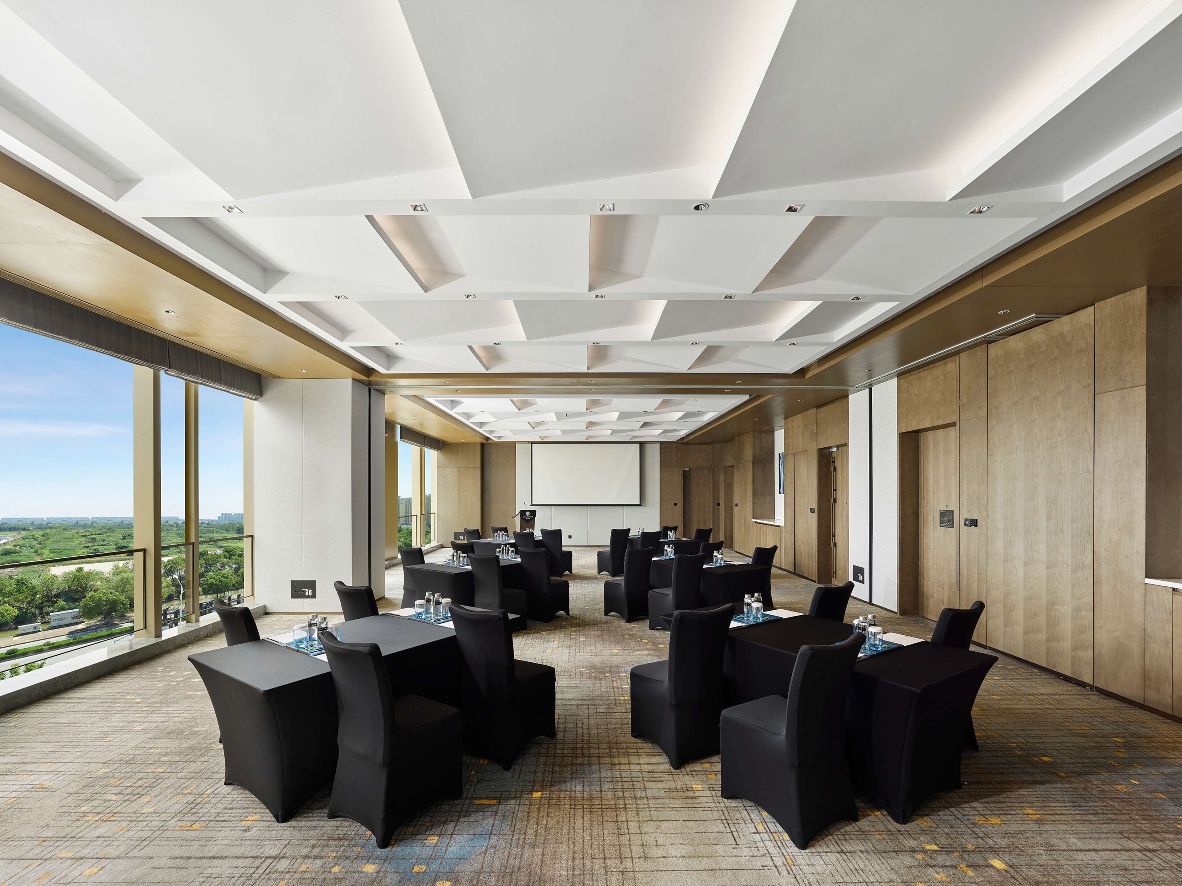 A conference room with multiple tables and chairs, large windows offering a view of the city, and a projector screen on the wall.