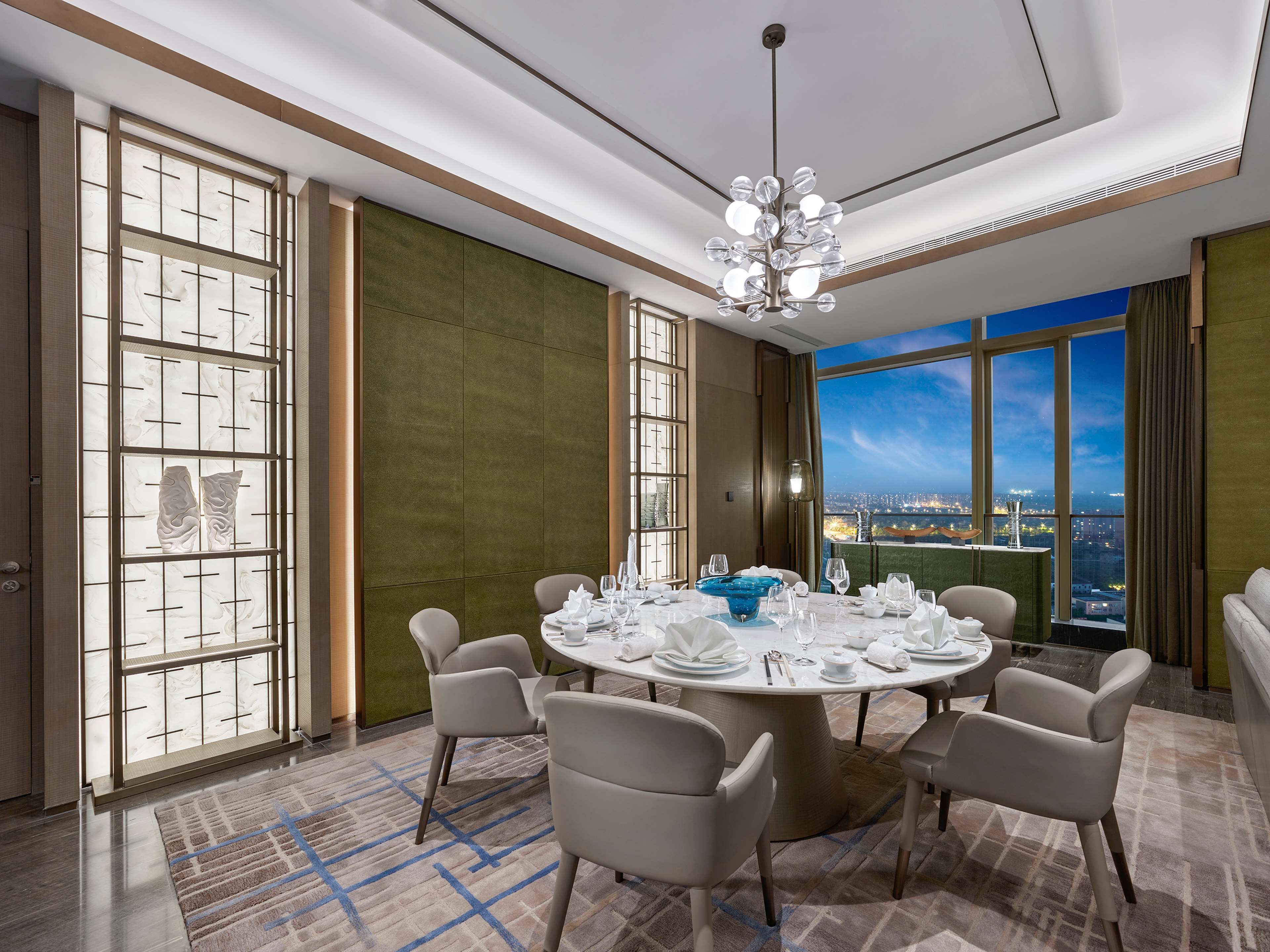 A modern dining room with a round table set for six, large windows with a city view, and a decorative ceiling light.