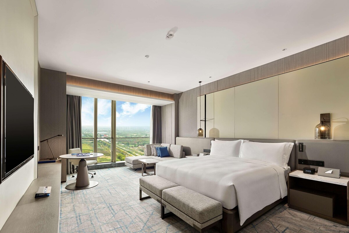 A modern hotel room with a large bed, a seating area, a desk, and a large window offering a city view.