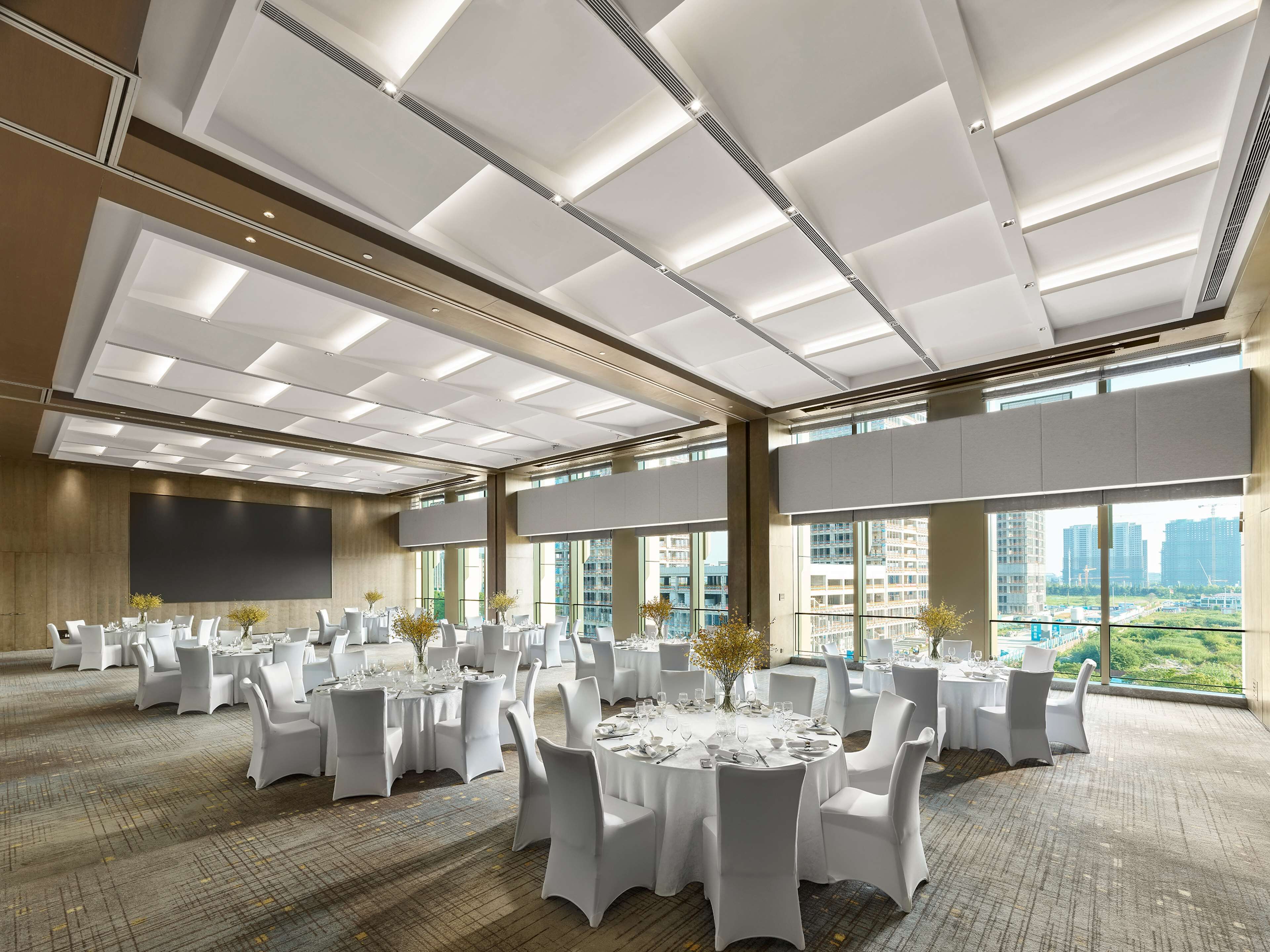 A spacious conference room with multiple round tables, white chairs, and a large screen on the wall.