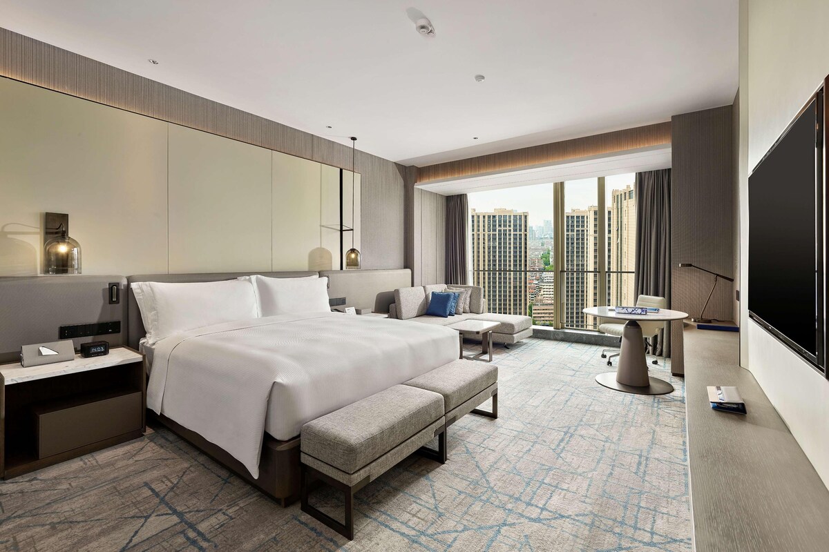 A modern hotel room with a large bed, a seating area, a small table, a TV, and a balcony with a city view.