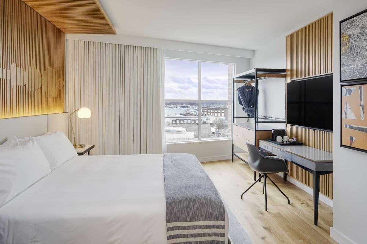 A modern hotel room with a large bed, a desk, and a view of the city through a large window.