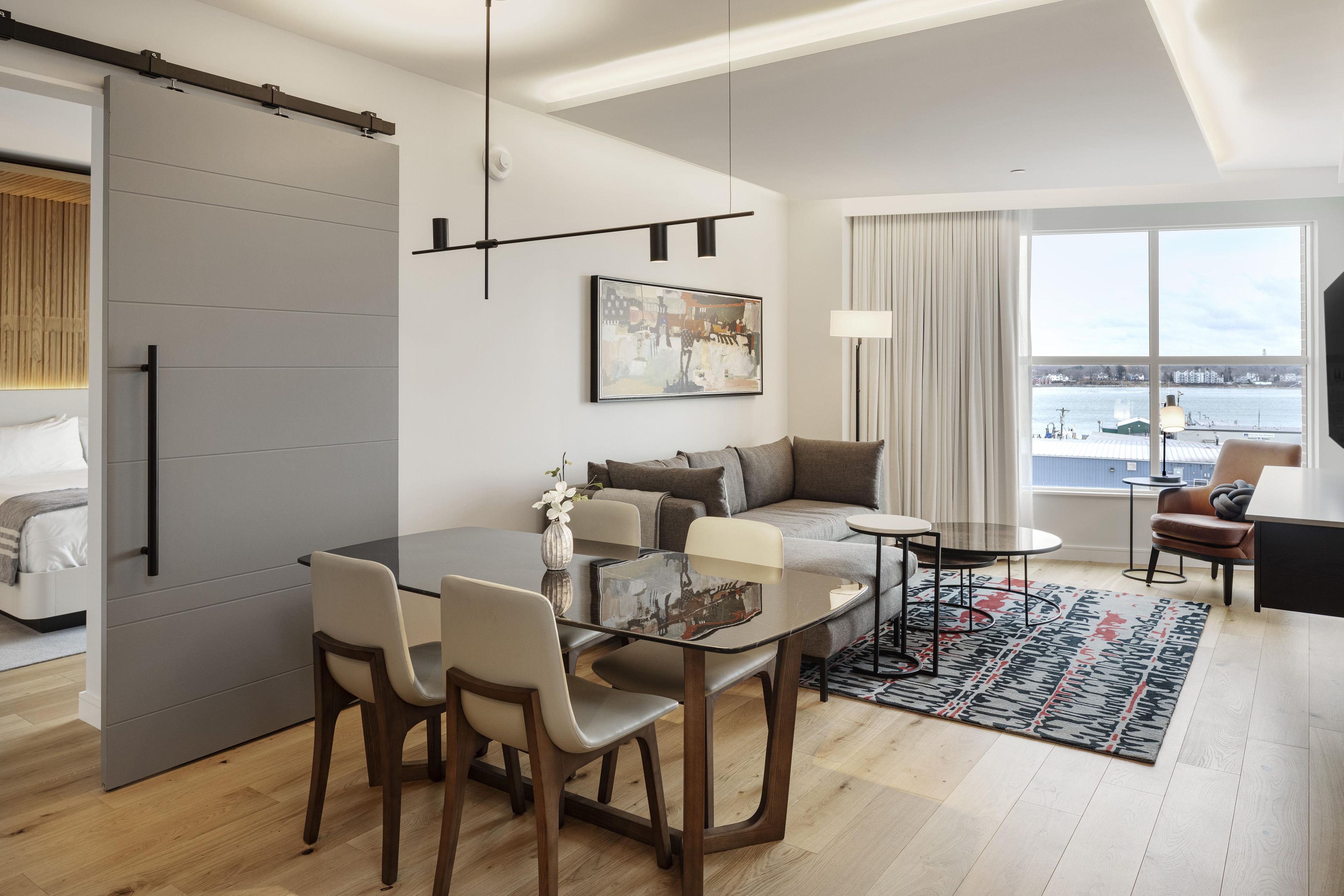 A modern living room with a sofa, a dining table with chairs, a large window with a view, and a sliding door.