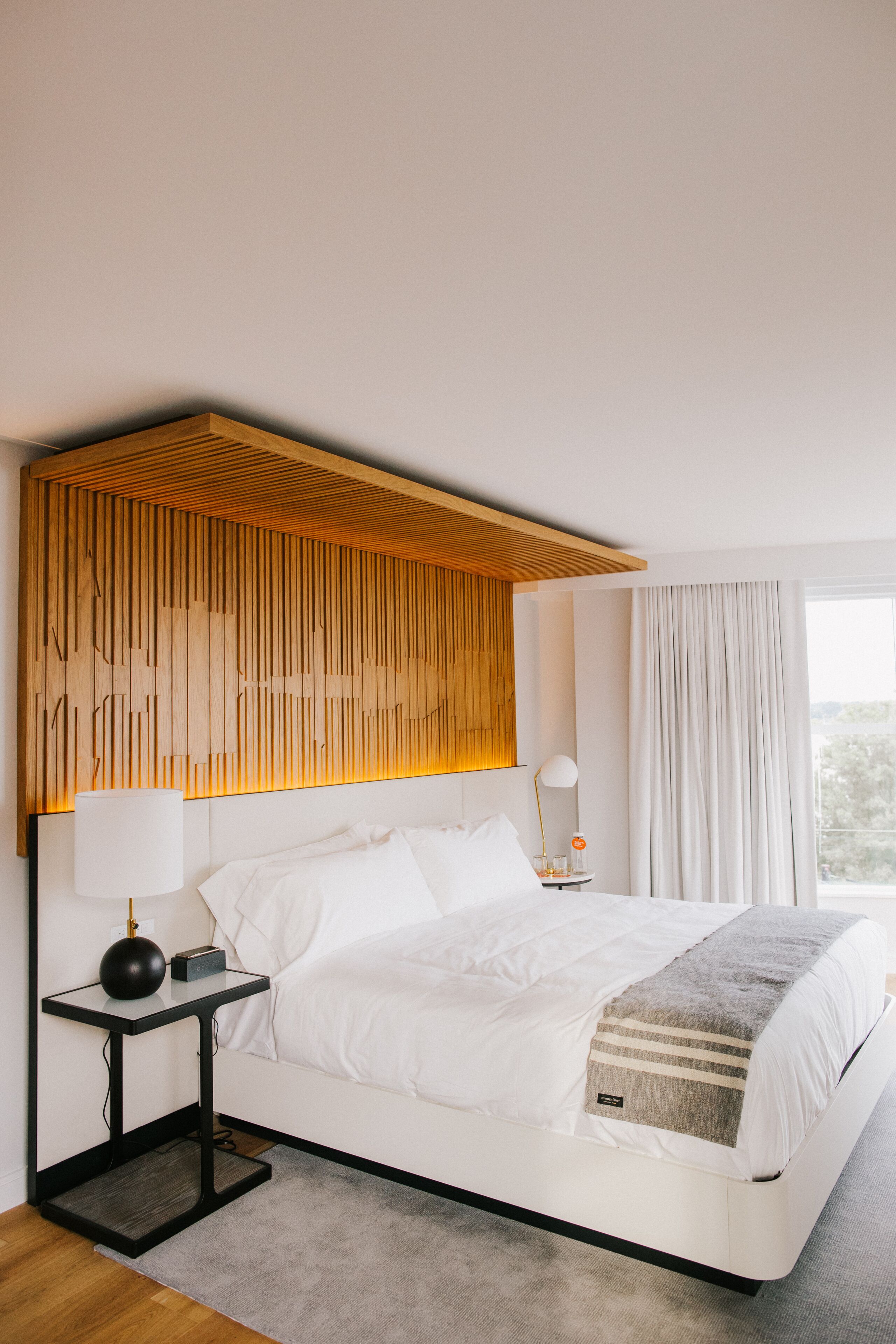 Une chambre d’hôtel moderne avec un grand lit, une tête de lit en bois, une table de chevet avec une lampe et une vue sur l’extérieur par une fenêtre.