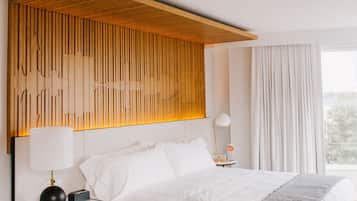 Une chambre d’hôtel moderne avec un grand lit, une tête de lit en bois, une table de chevet avec une lampe et une vue sur l’extérieur par une fenêtre.