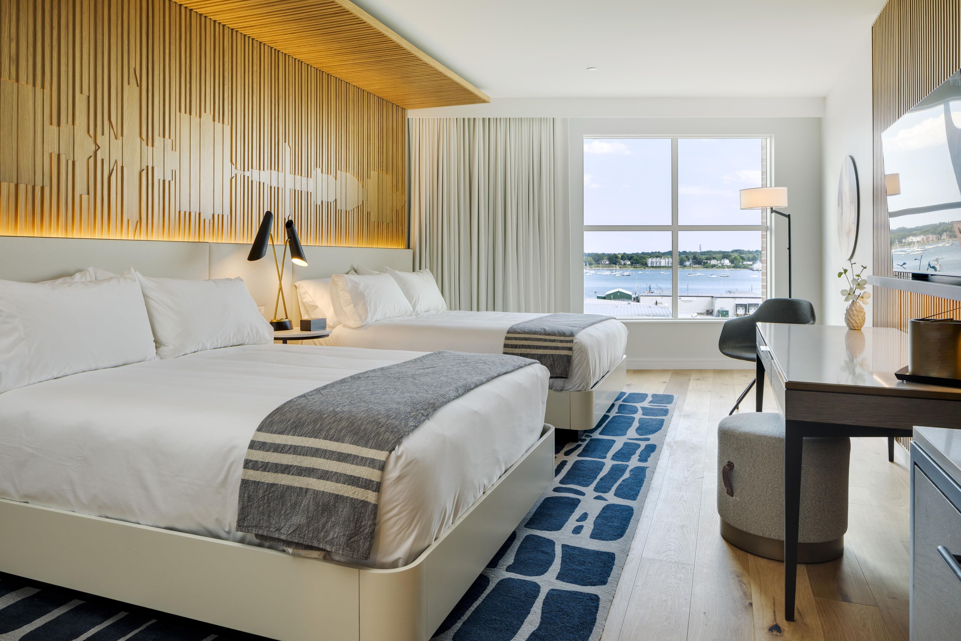 Une chambre d’hôtel avec deux lits, un bureau, une chaise, une grande fenêtre donnant sur la mer et un design moderne au niveau du plafond.