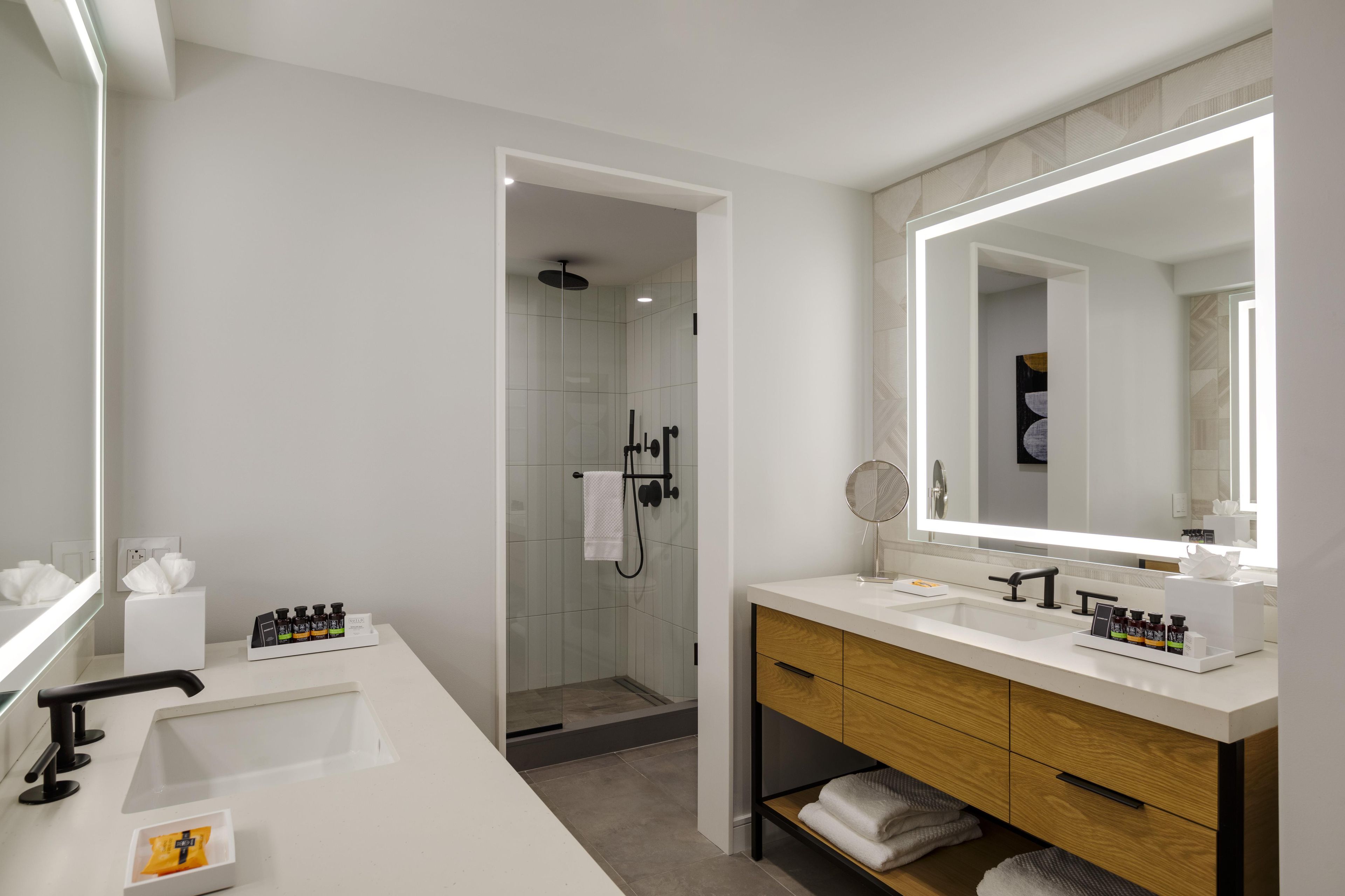 A modern bathroom with a large mirror, two sinks, a shower, and a vanity with storage.